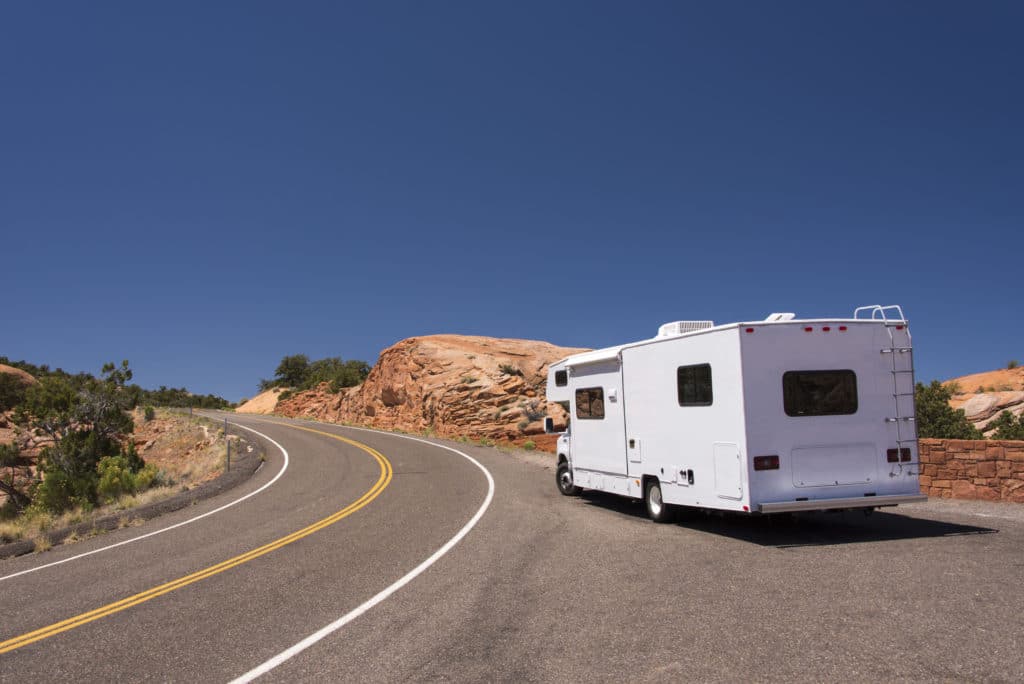 A white RV drives on a winding road through a desert landscape with rock formations under a clear blue sky.