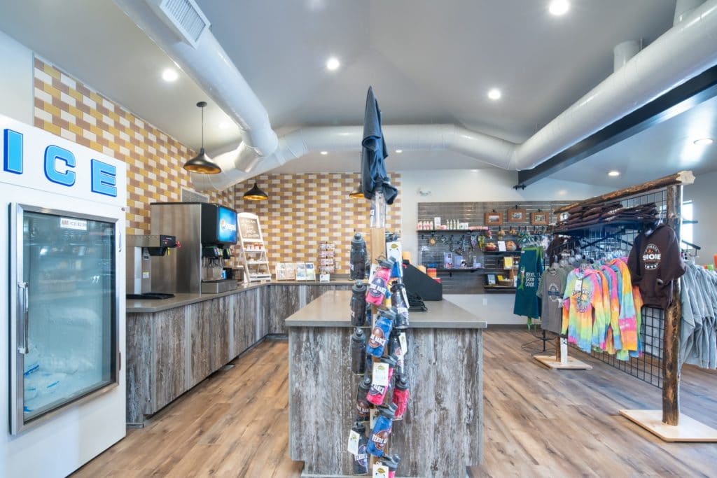 A convenience store interior with an ice freezer, beverage machines, clothing racks, and various items displayed on shelves and counters.