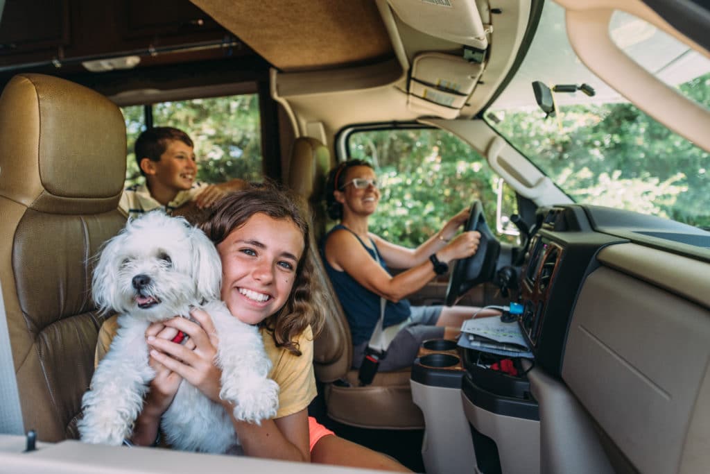 A girl holding a small white dog sits in the passenger seat of a vehicle, with a woman driving and a boy sitting behind them, all appearing happy.