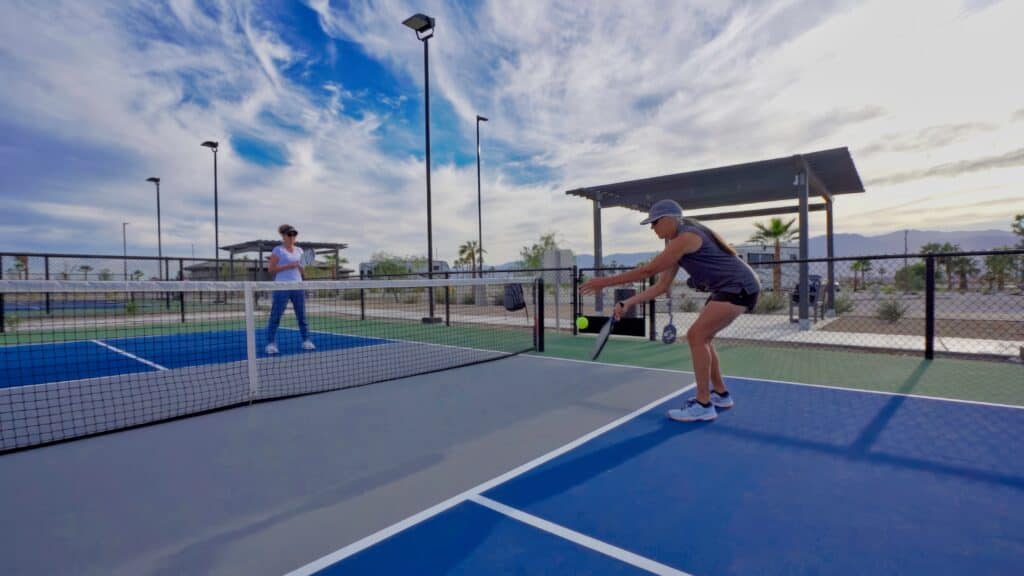Two people play pickleball on an outdoor court under a partly cloudy sky, with one person preparing to hit the ball near the net.