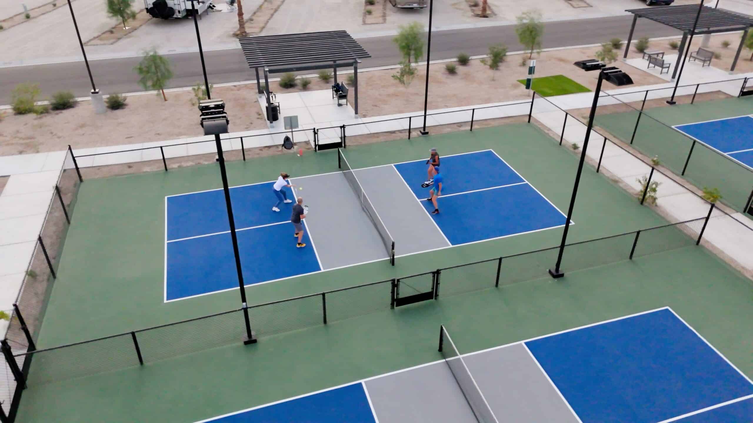 Aerial view of two pairs of people playing doubles on adjacent outdoor pickleball courts, surrounded by fencing and desert landscaping.