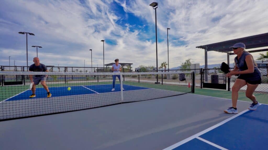 Three people are playing pickleball on an outdoor court under a partly cloudy sky, with one person preparing to hit the ball over the net.
