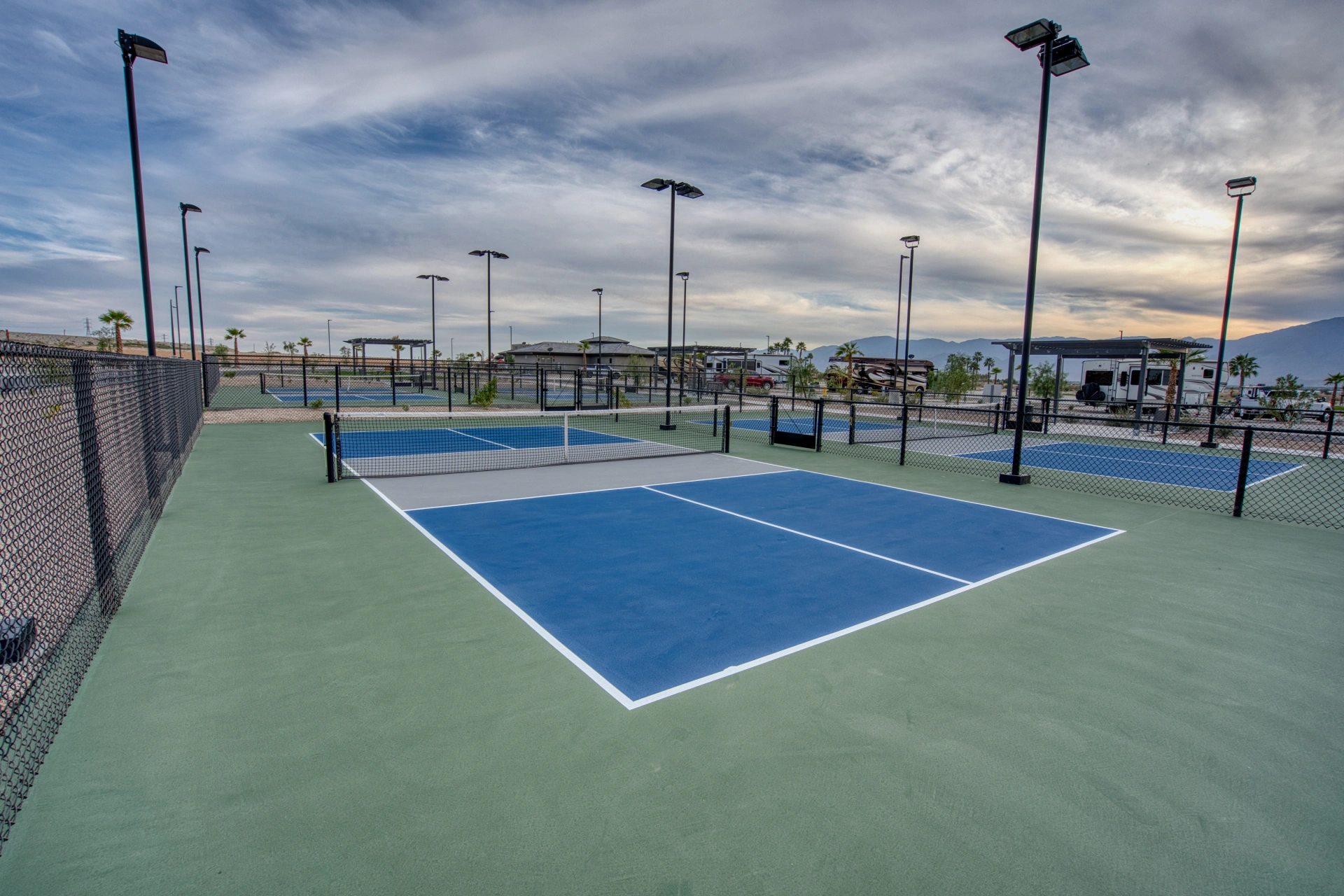 Outdoor pickleball courts with blue and green surfaces, surrounded by chain-link fencing, light poles, and a cloudy sky in the background.