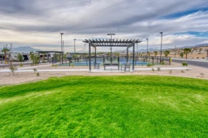 A shaded pergola with benches sits beside a tennis court, surrounded by landscaped grass, desert plants, and mountains in the background under a cloudy sky.
