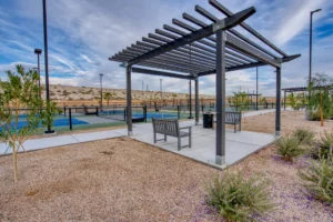 A shaded pergola with benches sits near fenced pickleball courts in a landscaped outdoor recreational area under a cloudy sky.