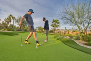 Two people on a putting green; one prepares to putt while the other steps near the hole. Desert landscaping and palm trees in the background.