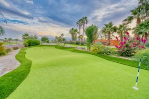 A manicured golf putting green is surrounded by palm trees, colorful flowers, and desert landscaping under a partly cloudy sky at sunset.