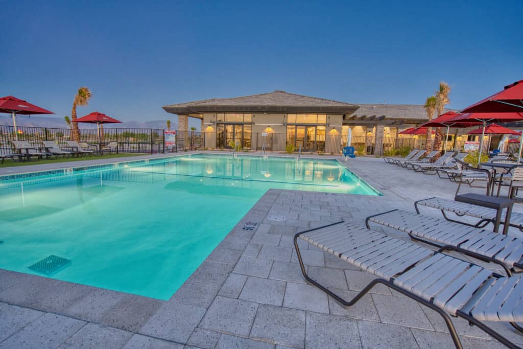 Outdoor swimming pool with lounge chairs and red umbrellas, adjacent to a modern building, photographed at dusk.