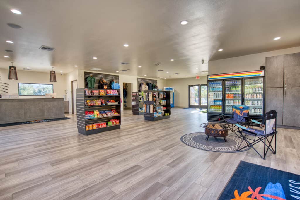 A spacious convenience store with shelves of snacks, drinks, and merchandise, a checkout counter, coolers along the wall, and folding chairs set up on a rug.