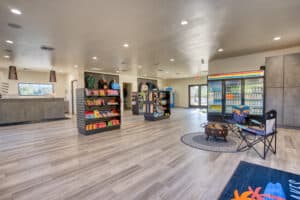 A spacious convenience store with shelves of snacks, drinks, and merchandise, a checkout counter, coolers along the wall, and folding chairs set up on a rug.