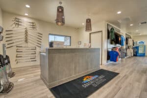Golf pro shop interior with a modern reception desk, golf clubs displayed on the wall, branded clothing on racks, and a logo mat on the floor.