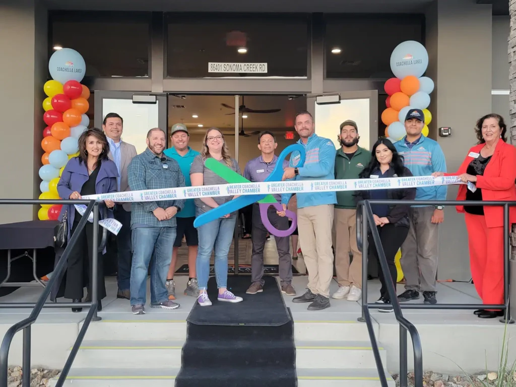 A group of people stand on steps in front of a building, holding a ribbon for a ribbon-cutting ceremony. Some hold large scissors; balloons decorate the entrance.