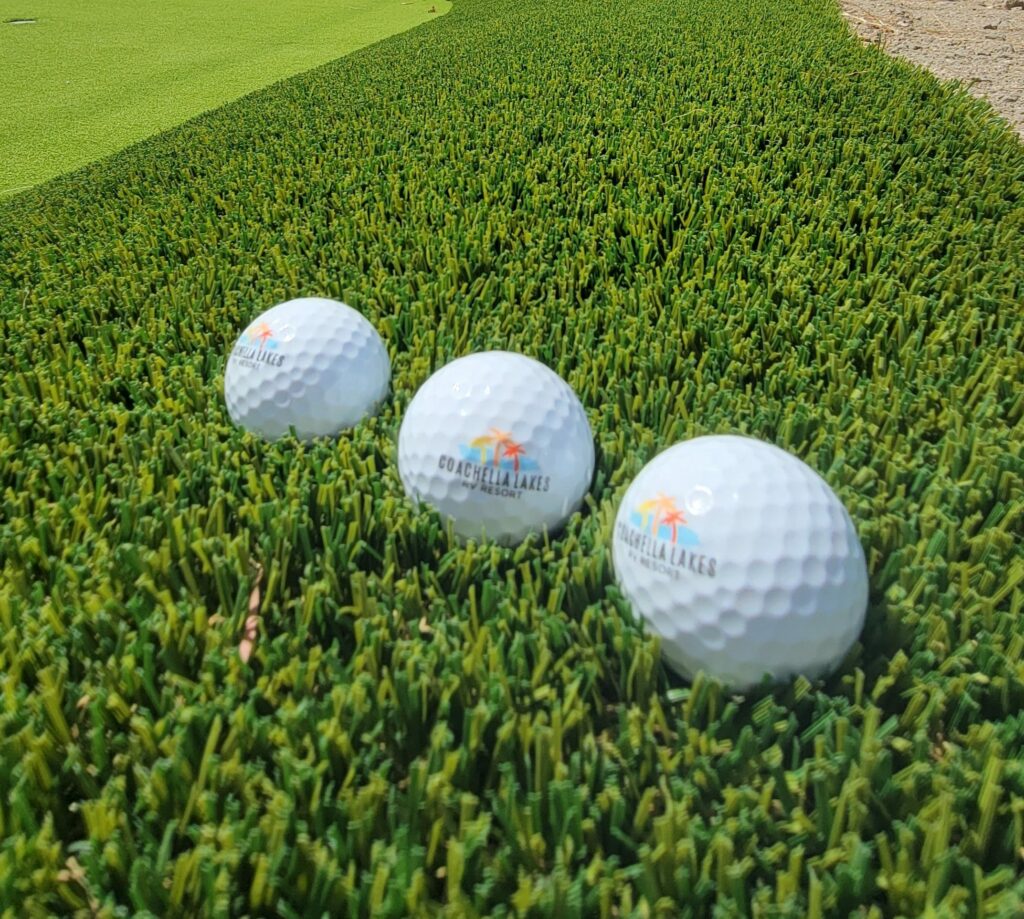 Three branded golf balls are lined up on artificial turf beside a putting green.