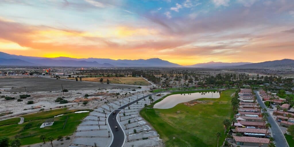 Aerial view of a suburban neighborhood with rows of houses, a small lake, parking lots, and distant mountains under a colorful sunset sky.