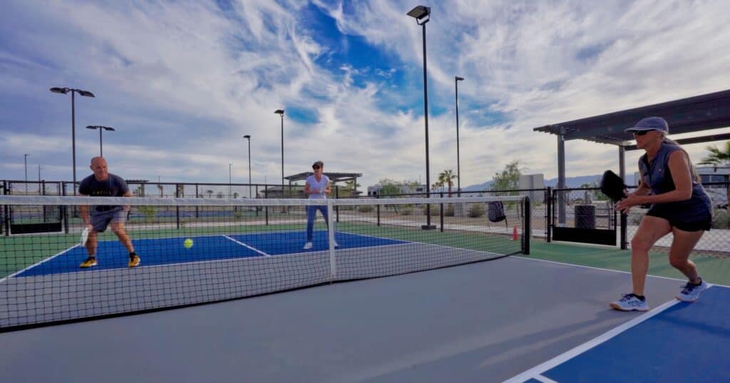Two people play pickleball on an outdoor court while a third person stands in the background. The sky is partly cloudy.