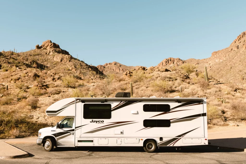 A white Jayco motorhome parked on pavement in front of a rocky desert landscape with cacti and clear blue sky.