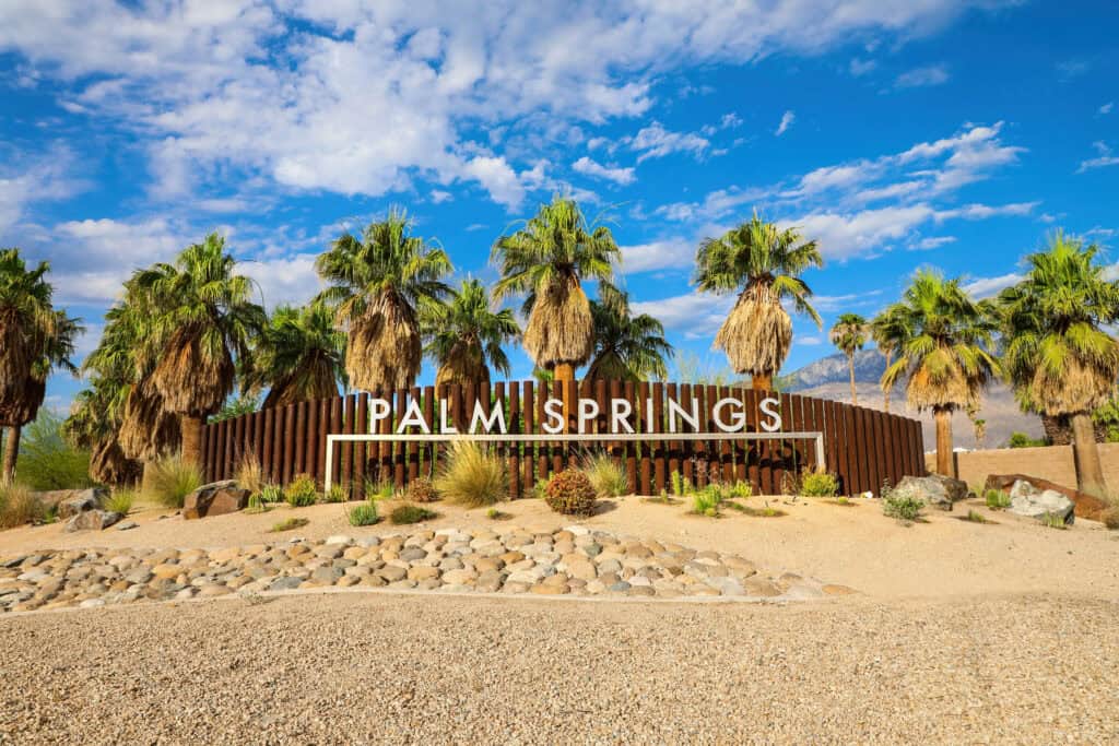 A large "Palm Springs" sign stands in front of palm trees, desert landscaping, and a blue sky with scattered clouds.