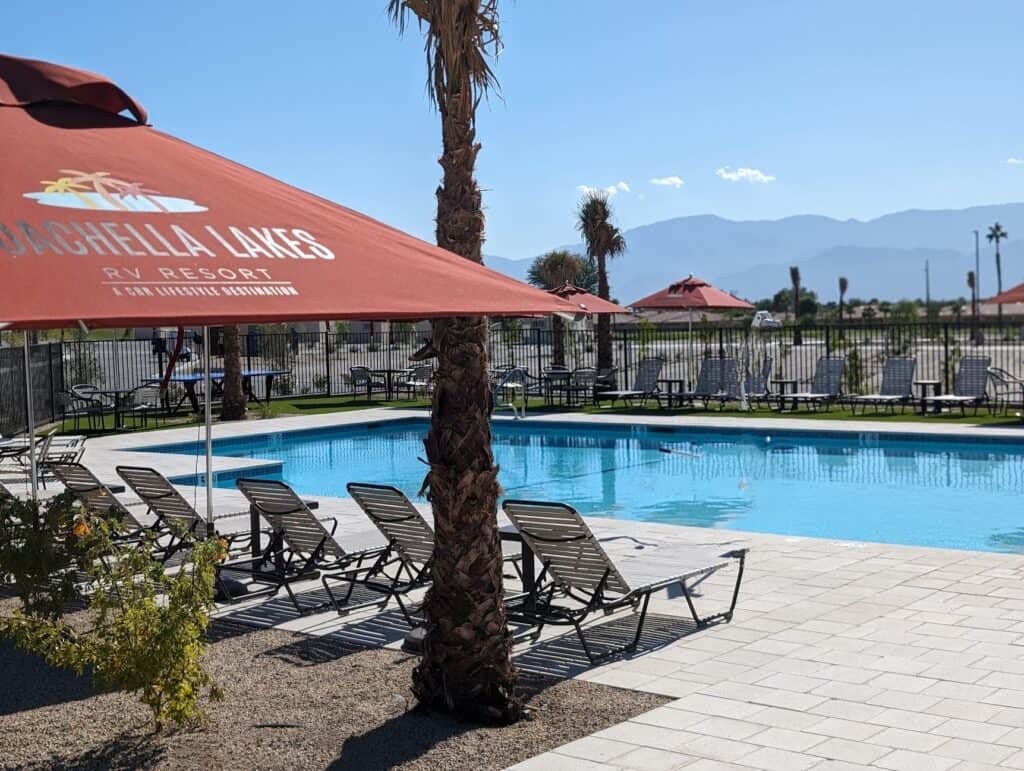 Outdoor swimming pool with lounge chairs and red umbrellas at Coachella Lakes RV Resort, with mountains visible in the background on a sunny day.