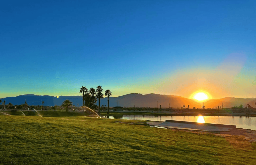 Sunset over distant mountains with a grassy park, palm trees, sprinklers running, and a calm body of water in the foreground.