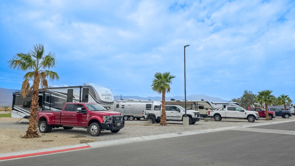Several RVs and pickup trucks are parked in a row at a desert campsite with palm trees under a partly cloudy sky.