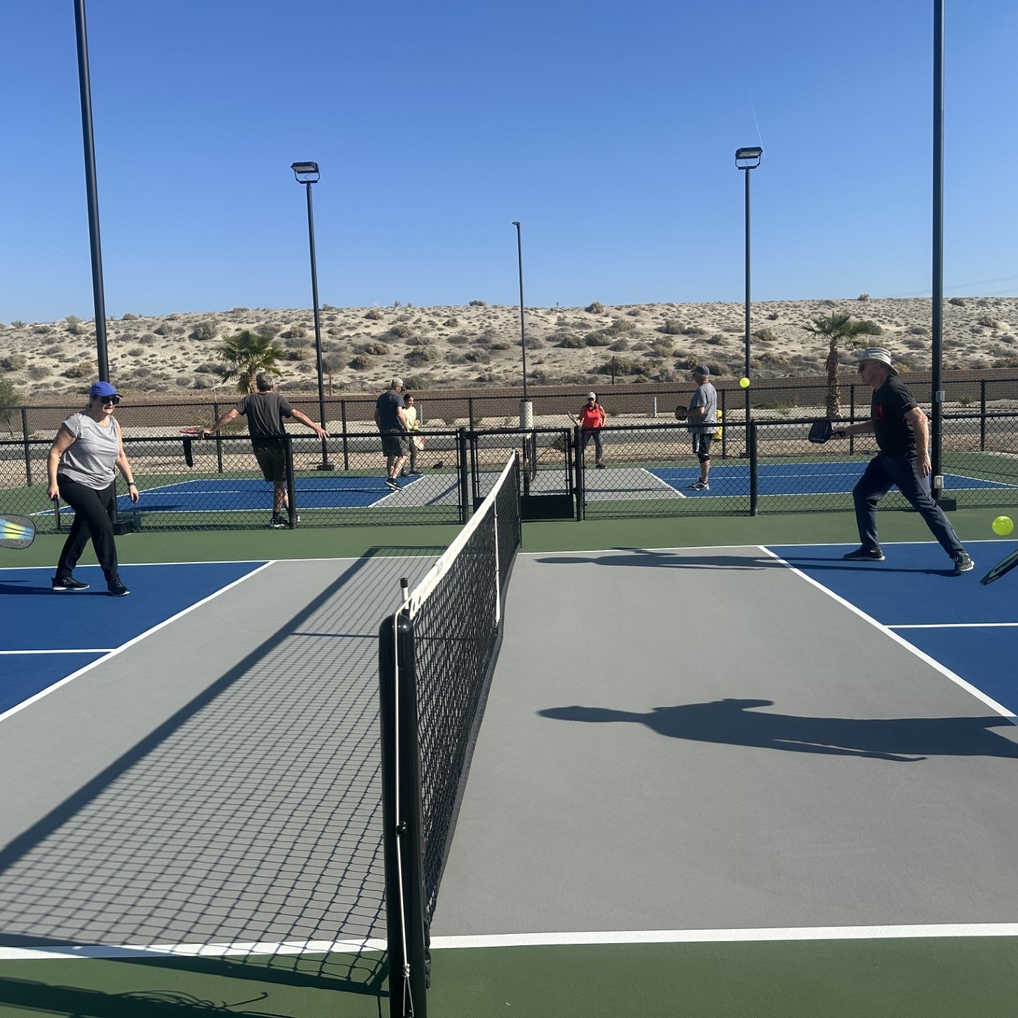 Six people are playing pickleball on outdoor courts under a clear blue sky, with a sandy landscape in the background.