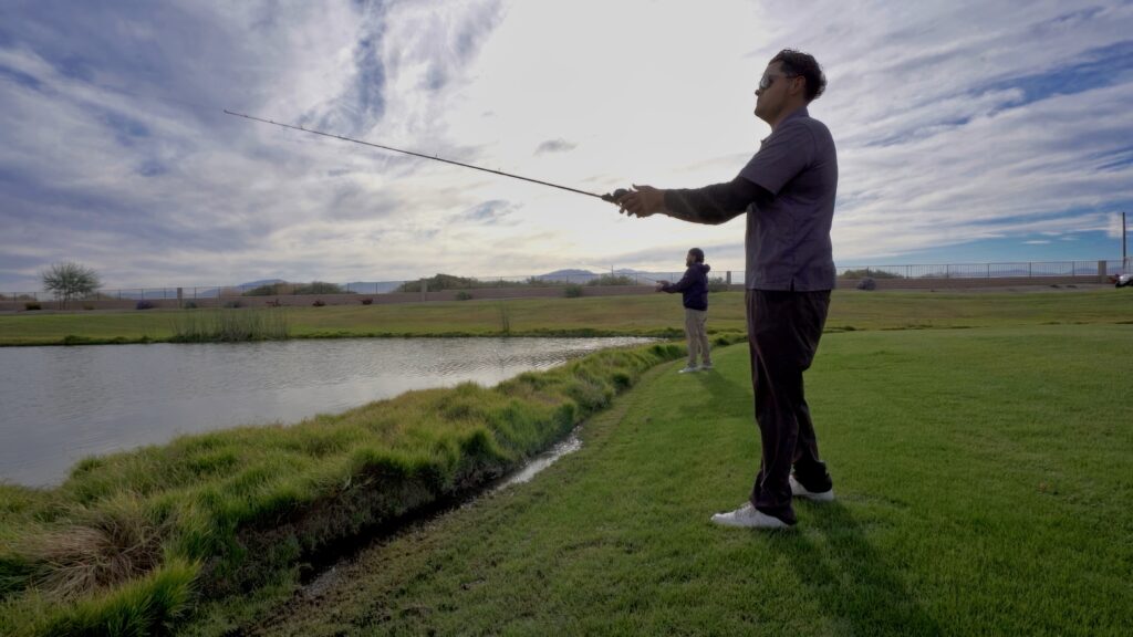 Two people stand on grassy ground next to a pond while fishing under a cloudy sky, with one person in the foreground and another in the distance.