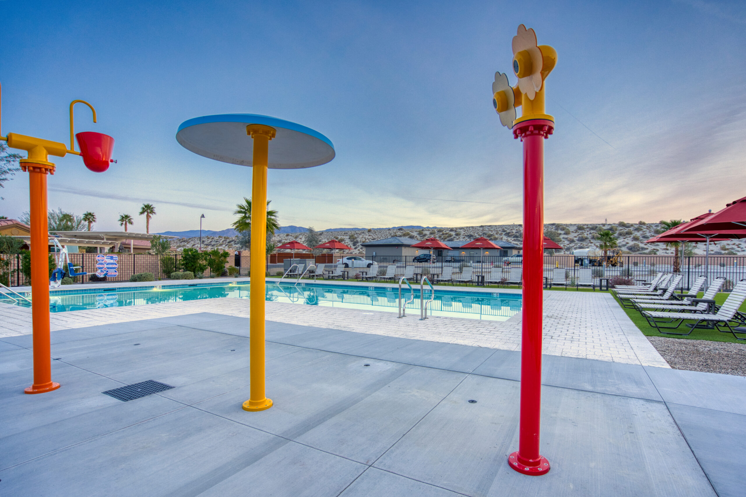 Outdoor swimming pool area with lounge chairs, umbrellas, and colorful splash pad features, set against a desert landscape under a clear sky.