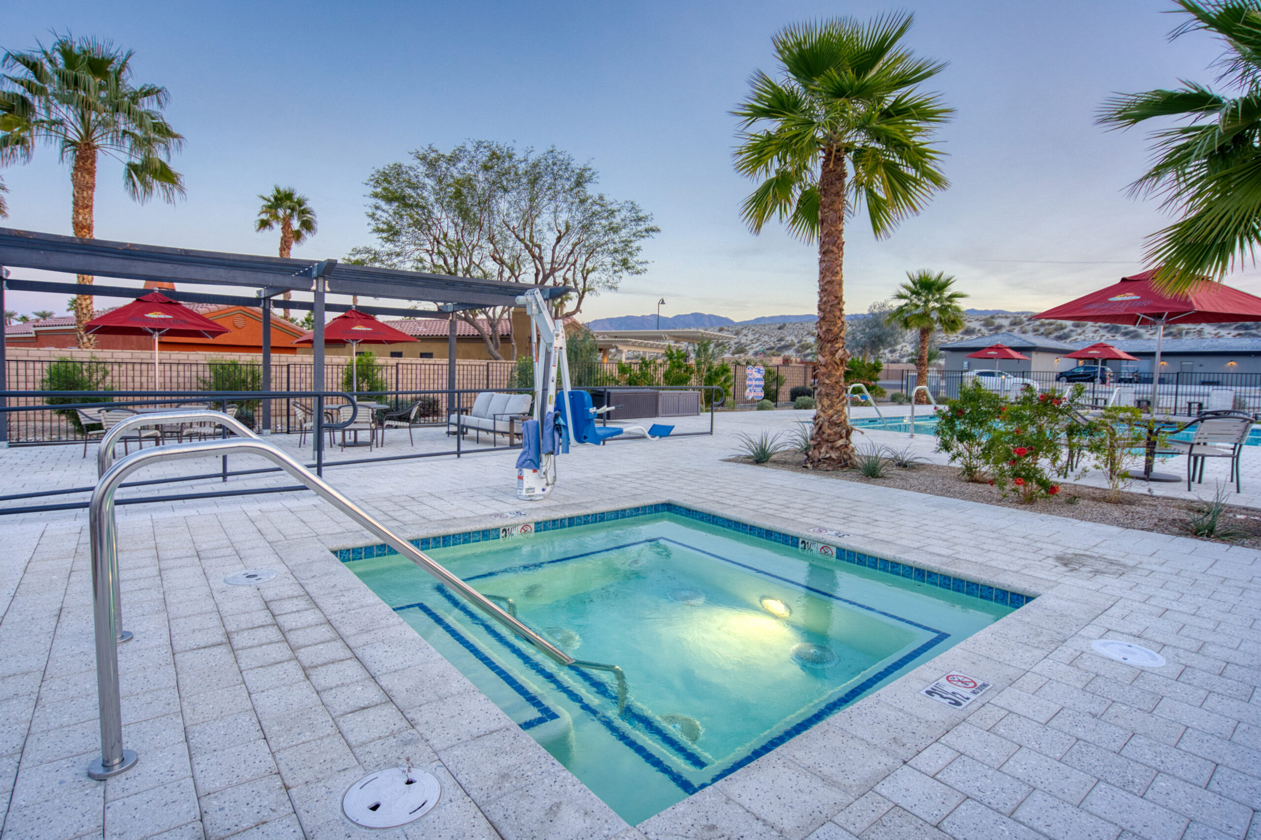 Outdoor hot tub with pool lift, surrounded by palm trees and lounge areas; a swimming pool and red umbrellas are visible in the background.