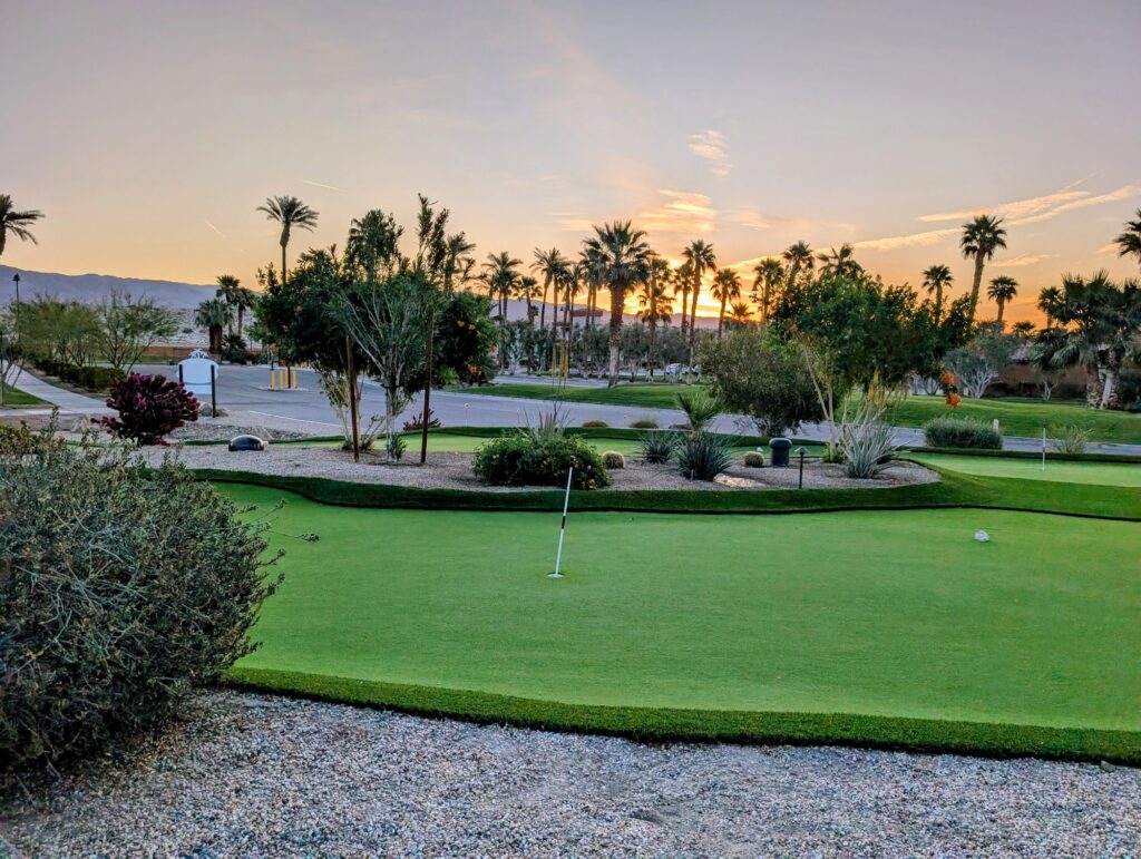A putting green surrounded by desert landscaping and palm trees at sunset, with mountains visible in the background.