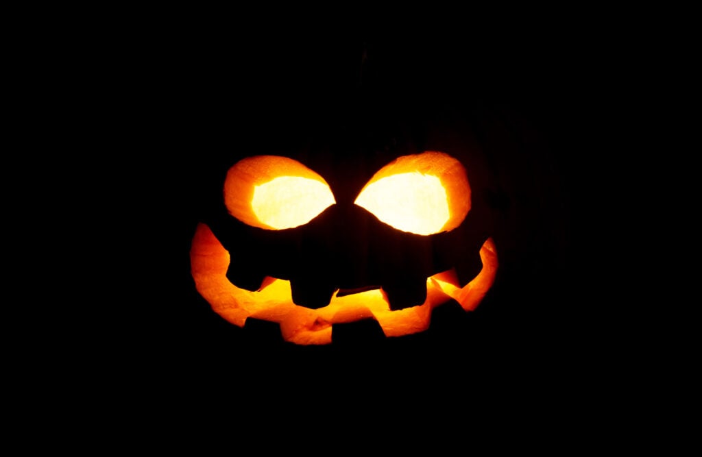 A carved pumpkin with glowing eyes and a jagged mouth is lit from inside, creating a jack-o'-lantern face against a dark background.