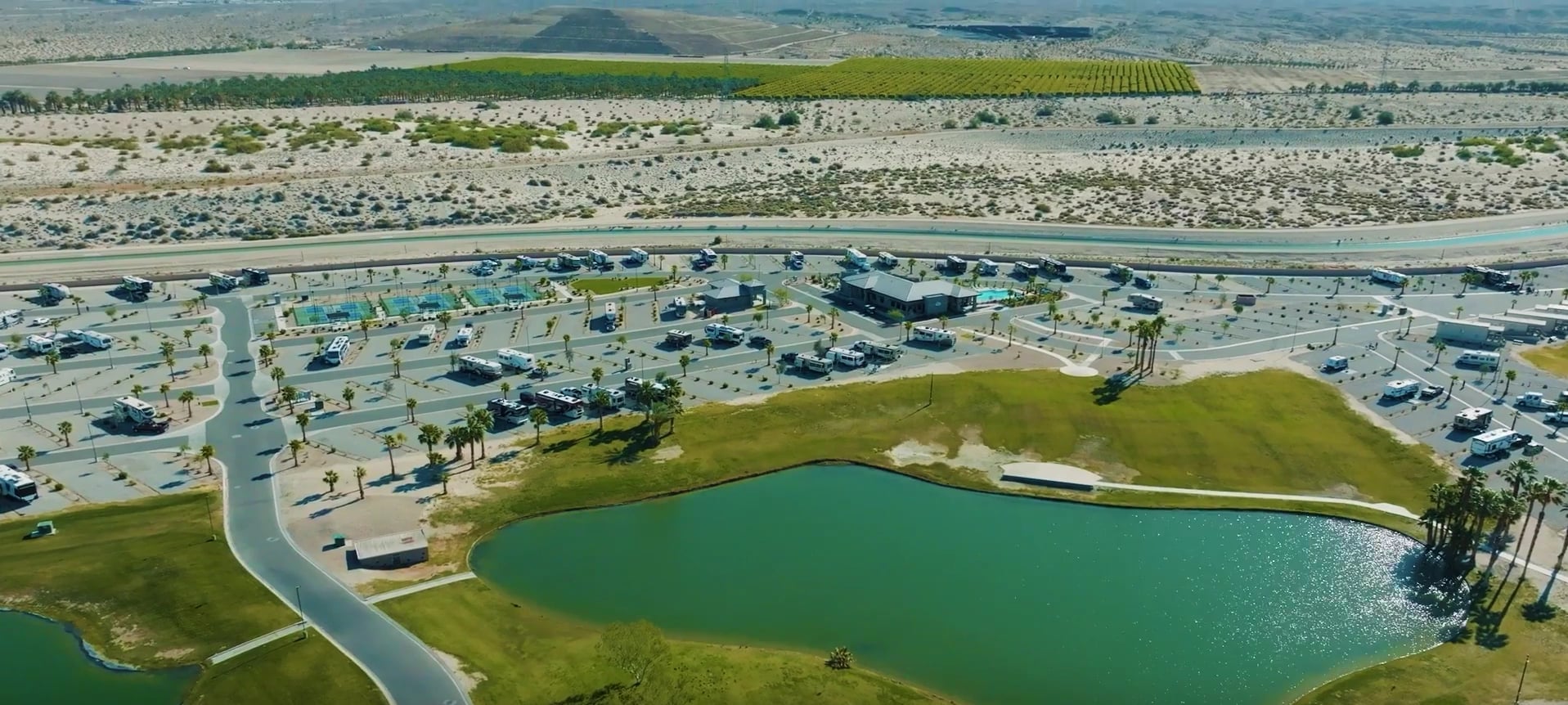 Aerial view of an RV park with numerous vehicles, green spaces, a pond, swimming pools, and desert landscape in the background.