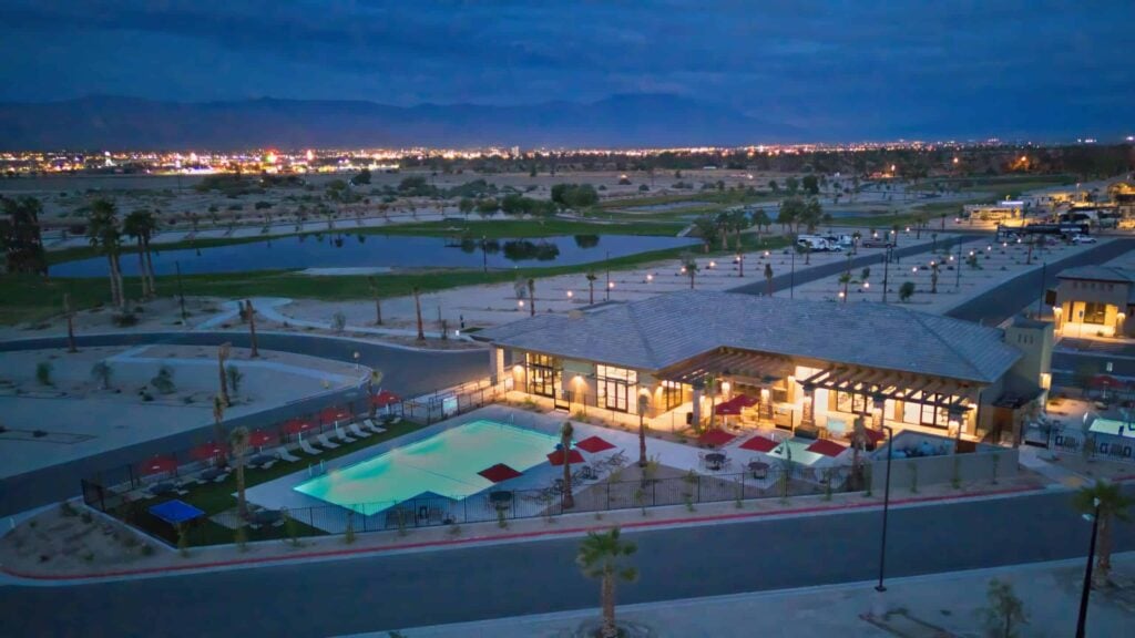 A brightly lit modern building with a swimming pool outside, seen at dusk, surrounded by a parking lot, palm trees, and distant city lights.