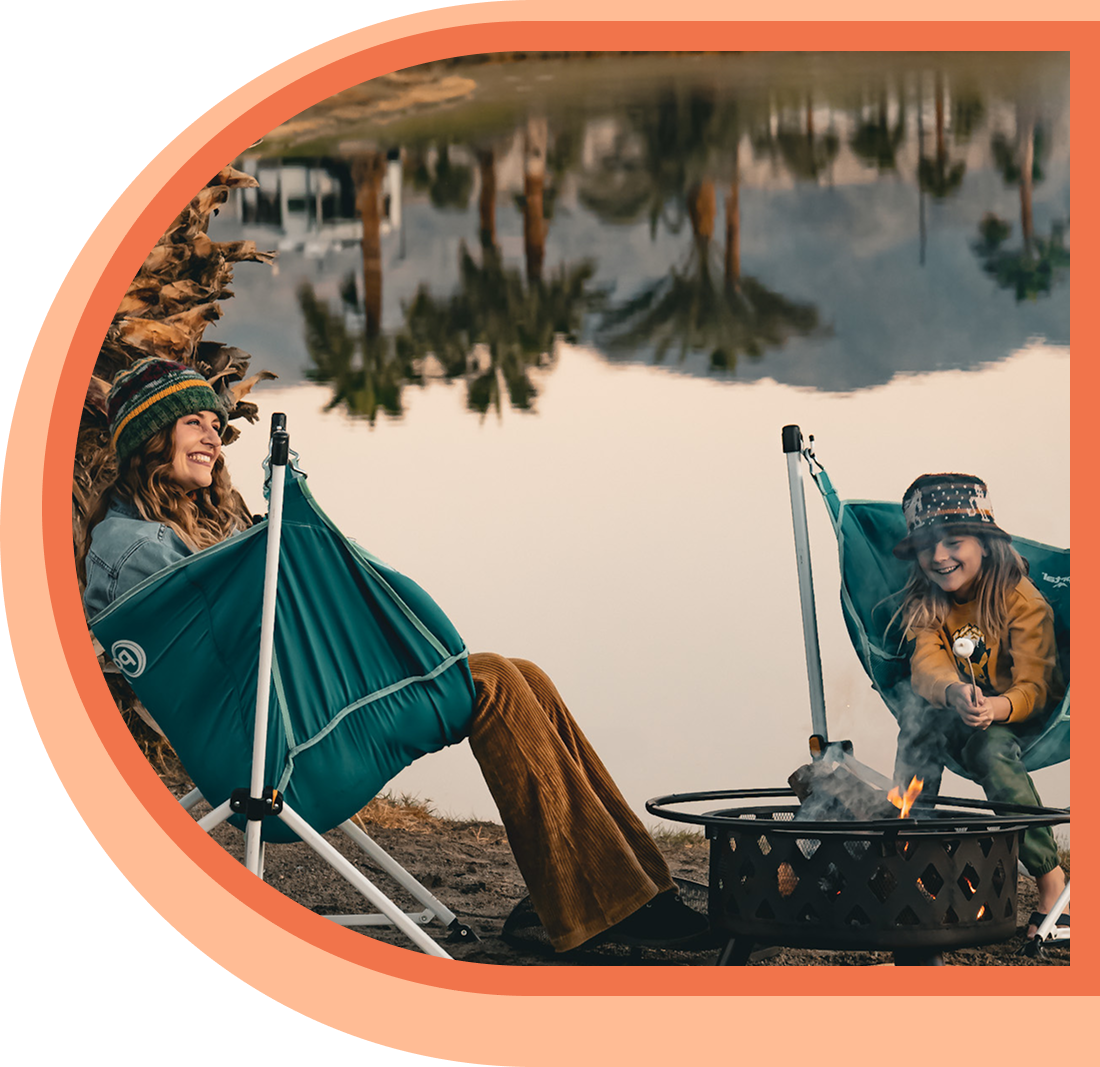 Two people sit in green camping chairs by a fire pit near a lake, roasting marshmallows, with trees and mountains reflected in the water.