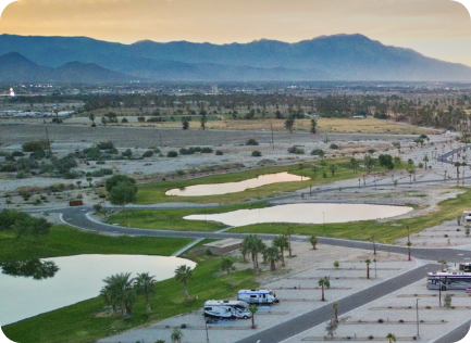 Aerial view of an RV park with several trailers, artificial ponds, desert landscape, scattered palm trees, and mountains in the background at sunset.