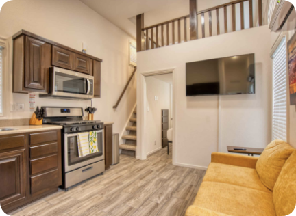 A small modern kitchen and living area with wood cabinets, stainless steel appliances, yellow sofa, wall-mounted TV, and stairs leading to a loft.