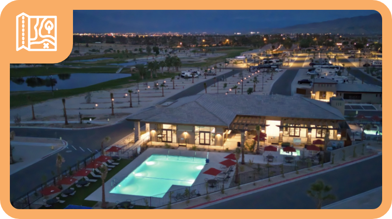 Aerial view of a modern building with a lit outdoor swimming pool, lounge chairs, and nearby RVs at dusk, with a cityscape visible in the distance.