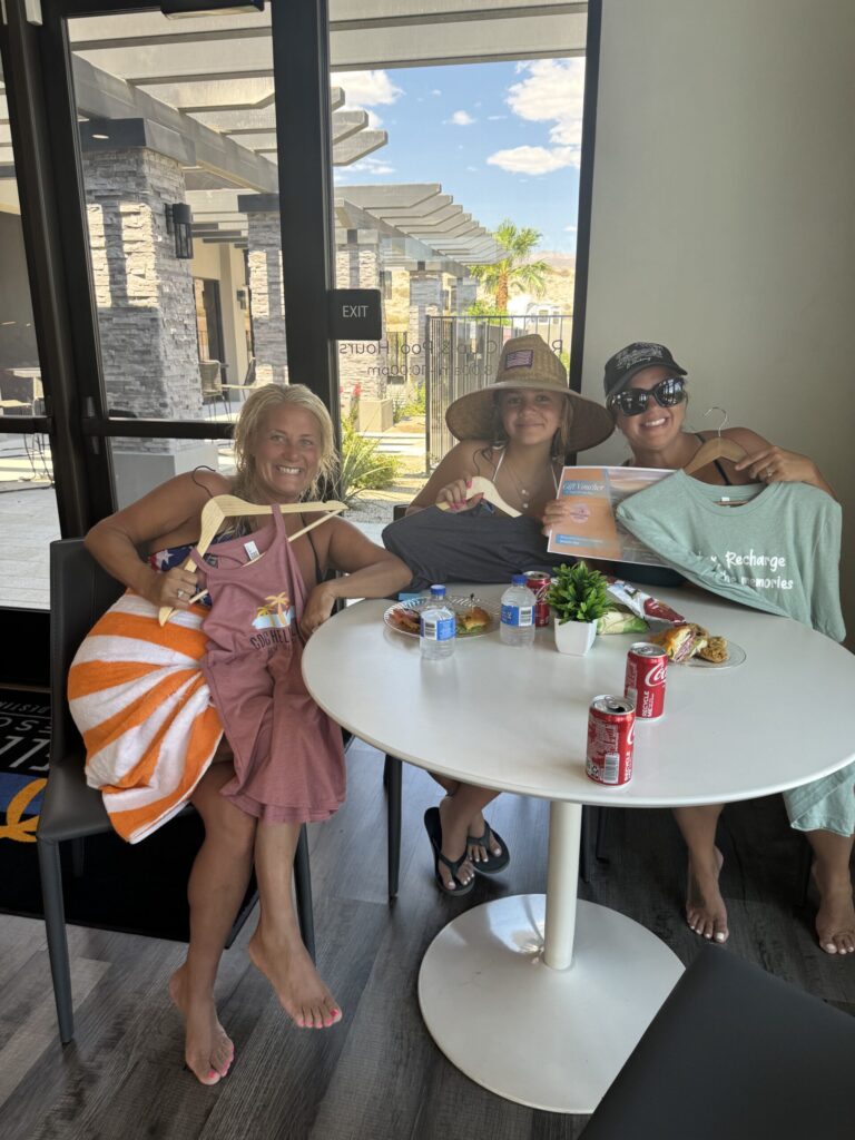 Three women sit around a round table with food, drinks, and clothing items. They are smiling, wearing swimsuits and sunhats, with towels and sunlight visible through the window behind them.