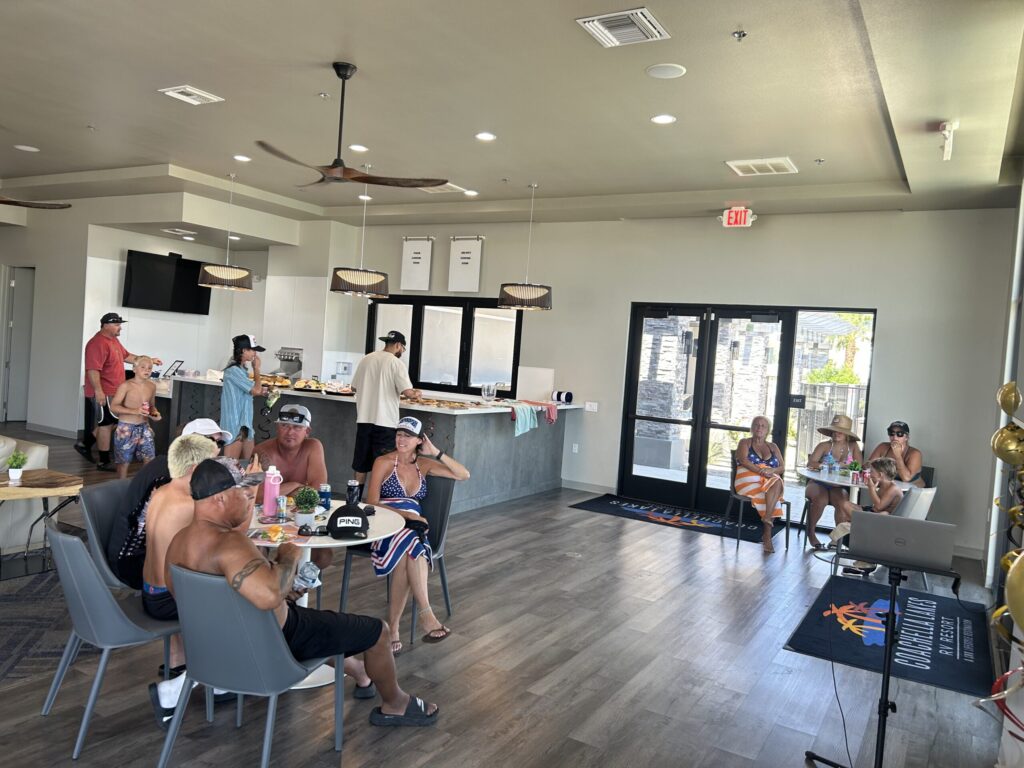 Several people sit and stand in a modern lounge area with tables, eating and socializing. The room has large windows, a TV, and overhead lights.