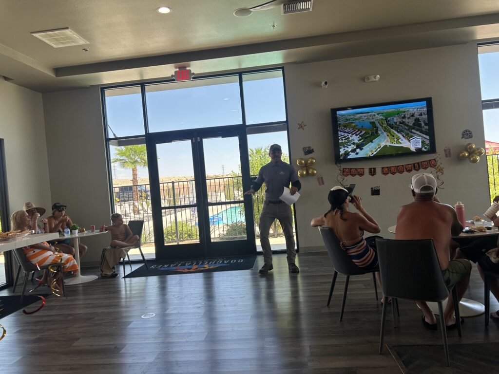 A man stands and speaks in front of a group of seated people in a room with a large window and a TV showing a community map; a pool is visible outside.