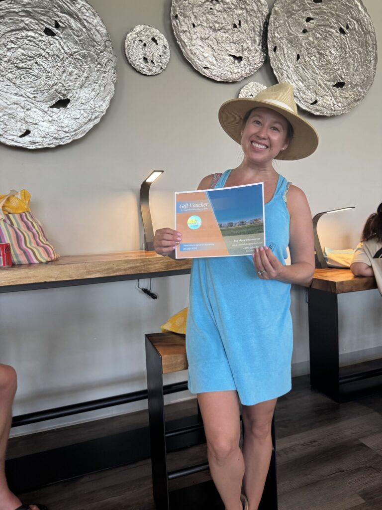 Woman in a blue dress and sunhat stands indoors, smiling and holding a certificate. Behind her, modern wall art and a counter with bags and seated people are visible.
