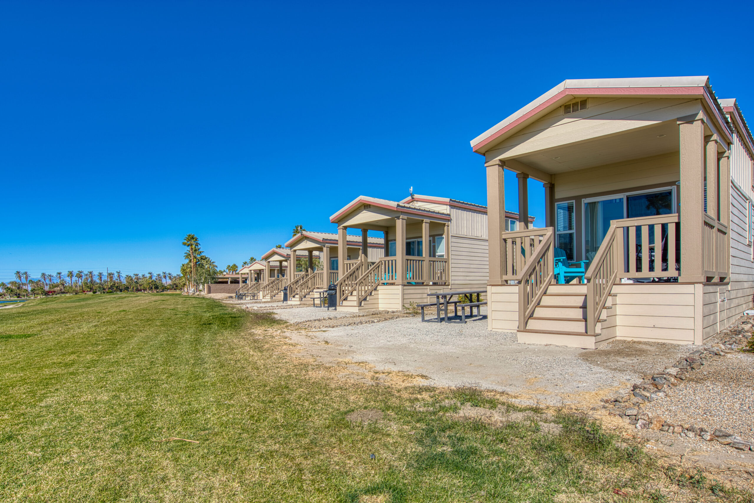 Row of small, modern cabins with porches facing a grassy area under a clear blue sky.