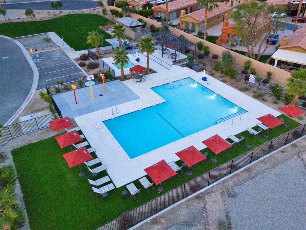 A rectangular outdoor swimming pool with red umbrellas, lounge chairs, a splash pad, and nearby parking in a fenced community area.