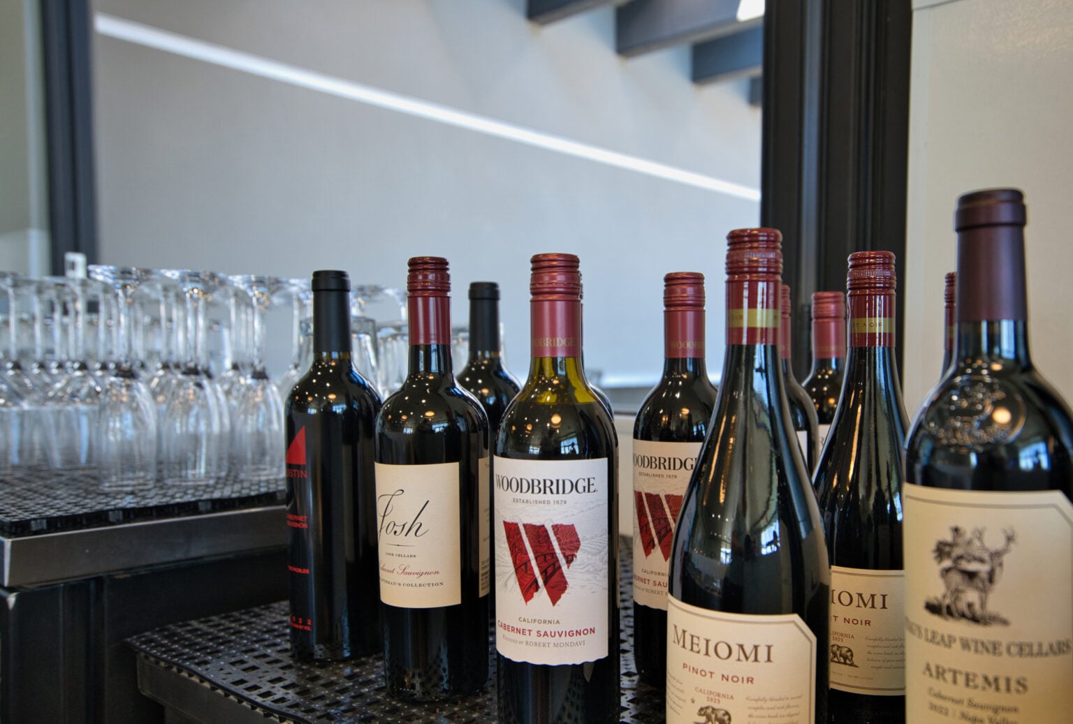 Several bottles of red wine are lined up on a black counter next to rows of empty glassware in a modern indoor setting.