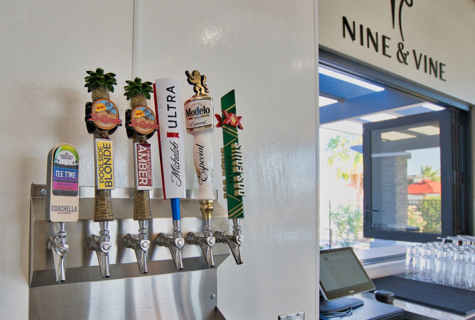 Six beer taps with branded handles are mounted on a wall beneath the Nine & Vine sign, next to a window and a counter with glasses and a computer terminal.
