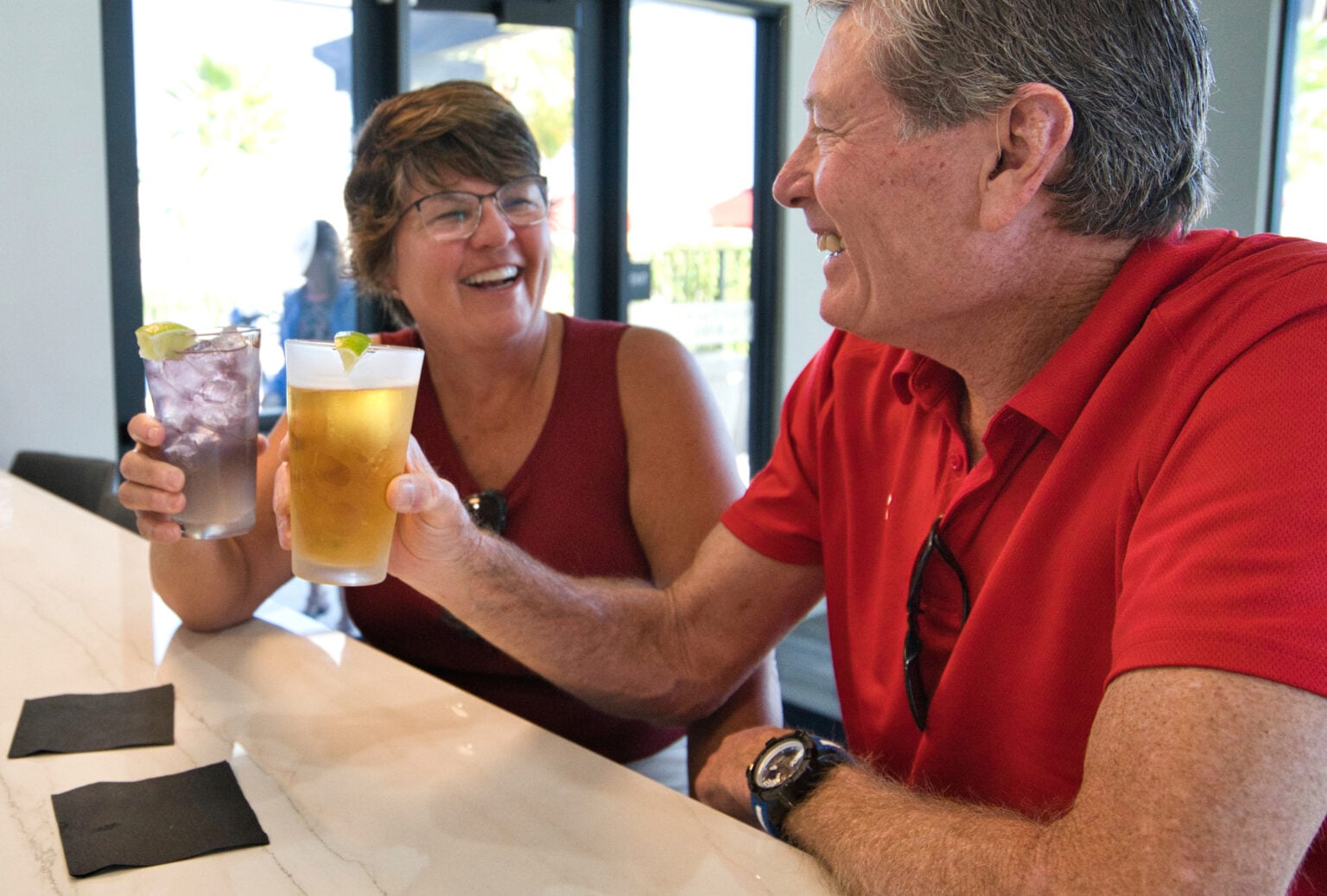 Two people sit at a bar, smiling and clinking their drinks, one with a beer and the other with a cocktail, both wearing red tops.