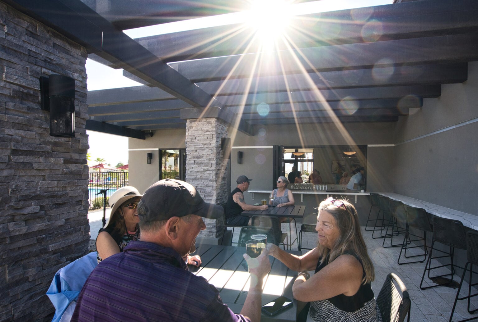 People sit and converse at outdoor tables under a pergola, with sunlight streaming through the beams.