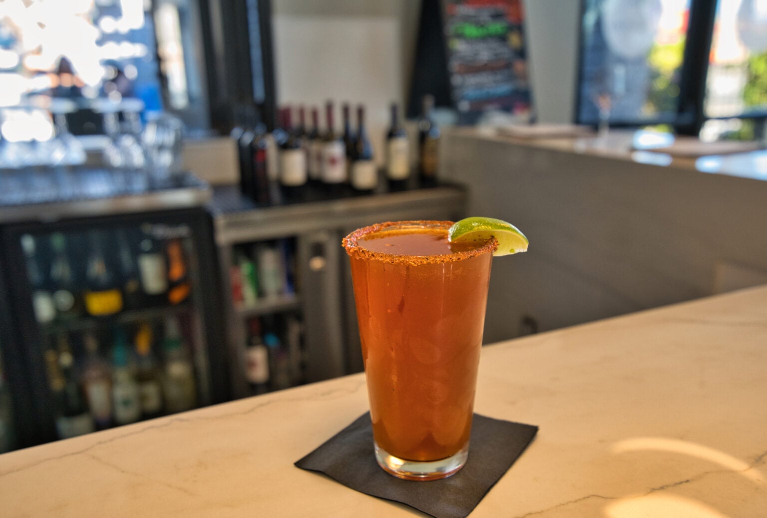 A glass of michelada with a chili powder rim and lime wedge sits on a black napkin at a bar counter, with bottles and a menu board in the background.
