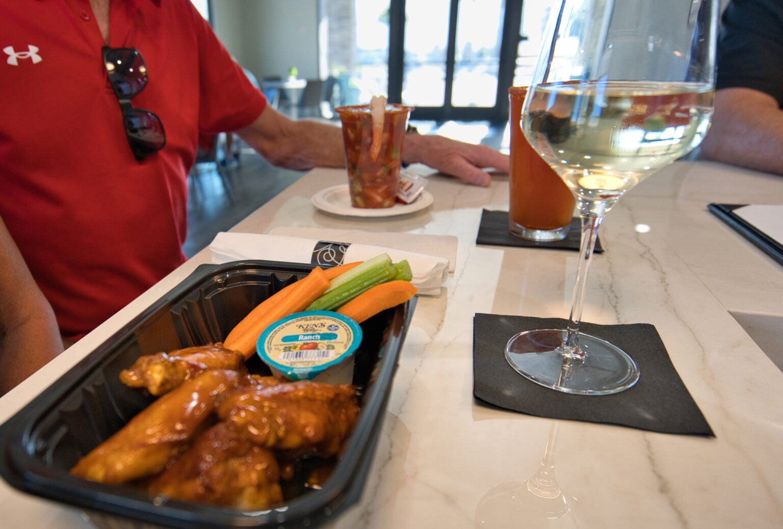 A tray of buffalo wings with celery, carrots, and blue cheese dip sits on a counter next to a glass of white wine and a Bloody Mary. A person in a red shirt is seated nearby.