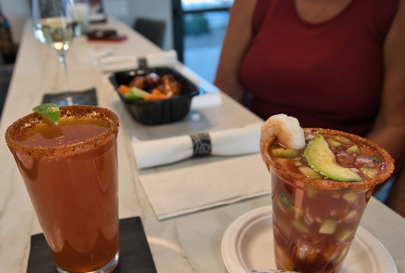 Two drinks with chili powder rims sit on a counter; one is garnished with lime, the other with shrimp and avocado. A takeout container and a person in a red shirt are in the background.