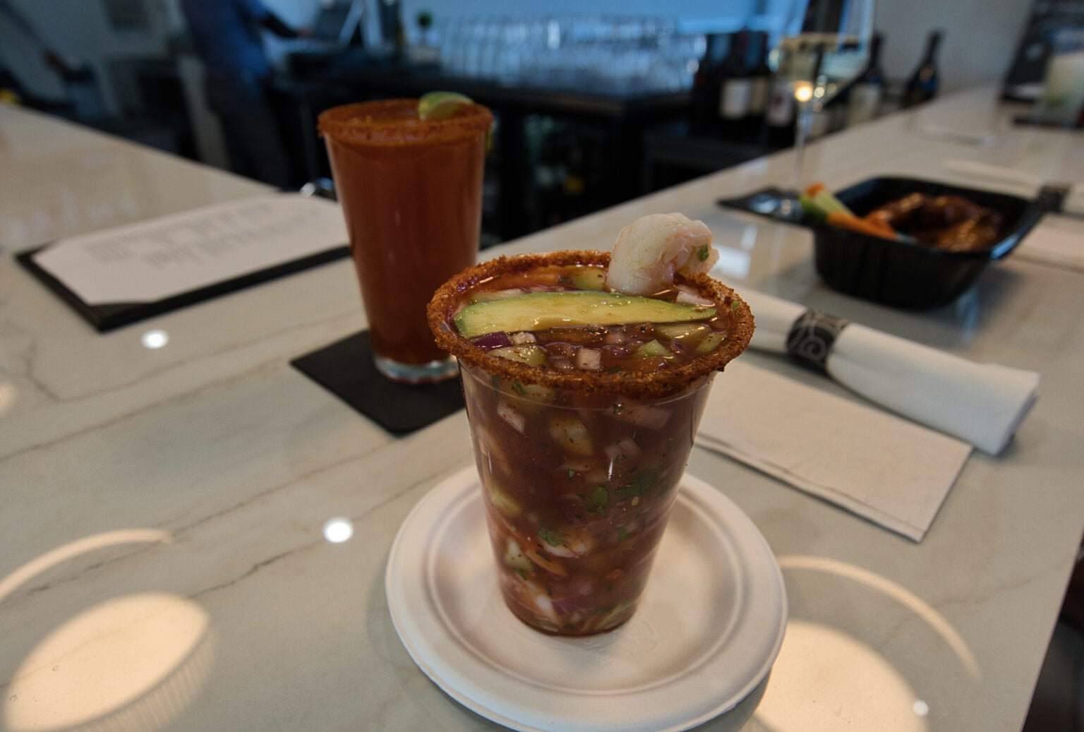 A glass of shrimp cocktail with avocado and a slice of cucumber, rimmed with chili powder, sits on a counter next to a drink and a takeout container.